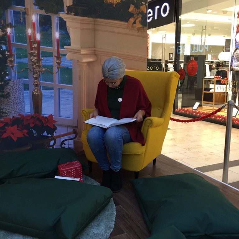 Ältere Dame in roter Jacke und Jeans, die auf einem gelben Sessel sitzt und aus einem Buch vorliest.
