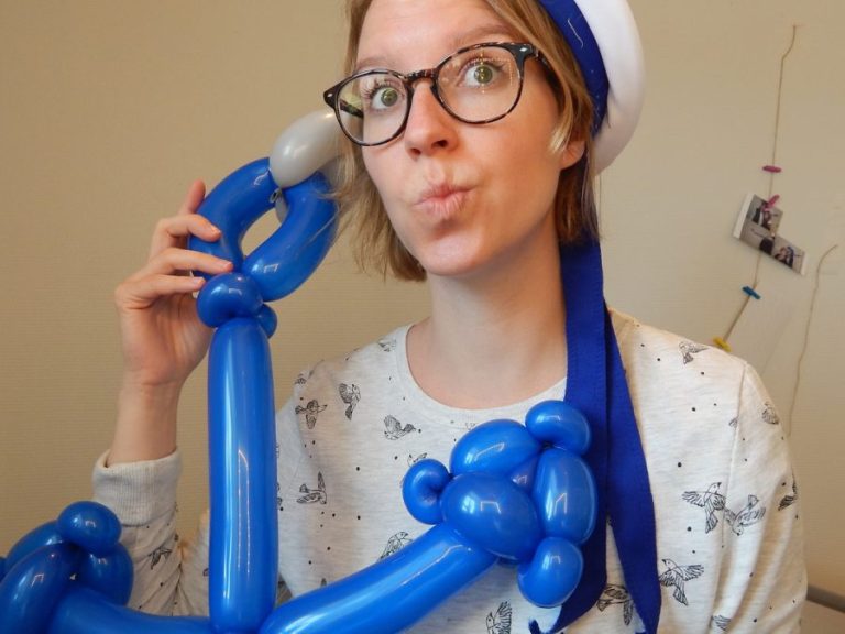 Frau mit weißem T-Shirt, großer Brille und einem weiß-blauen Semmanshut. In ihrer Hand hält sie ein blaues Ballontier.