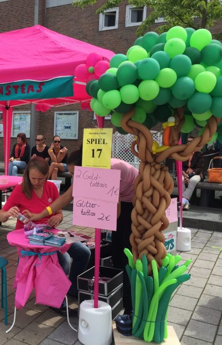 Frau an einem Straßenstand für Glitzertattoos. Neben dem Stand steh ein großer Ballonbaum.