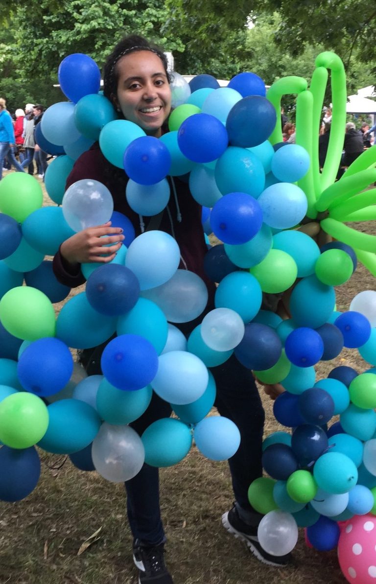 Eine lächelnde junge Frau, die am ganzen Körper mit blauen und grünen Ballons behängt ist.