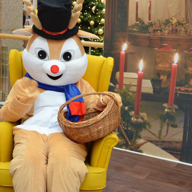 Person, die als Rentier-Maskottchen verkleidet ist. Sie sitzt auf einem gelben Sessel vor weihnachtlicher Dekoration.