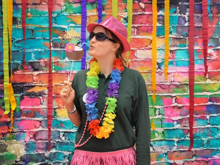 Frau in Hose und T-Shirt mit bunter Blumenkette, rosa Bastrock und rosa Hut. In ihrer Hand hält sie die Pappnachbildung eines Cocktails in einem Kokosnuss-Glas.