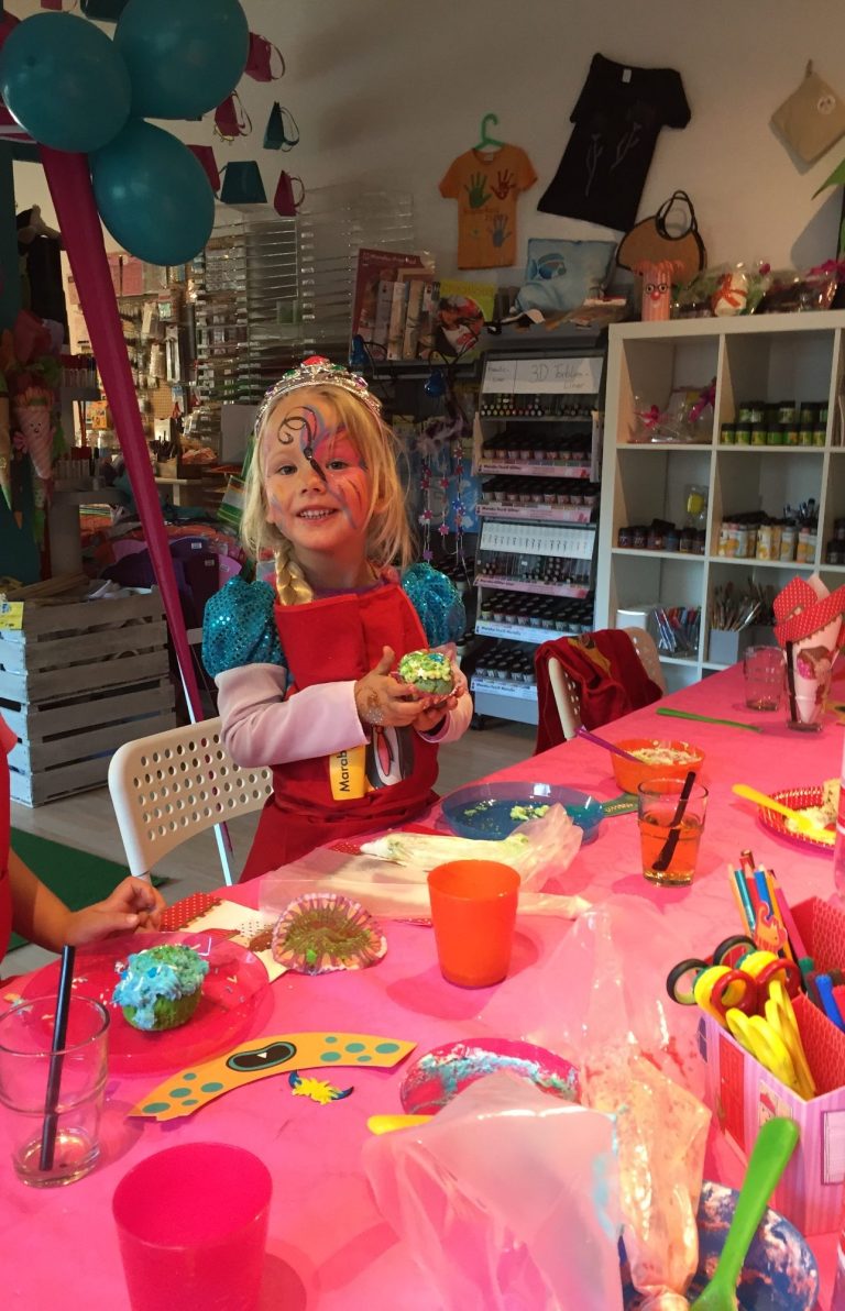 Blondes Mädchen mit Prinzessinnenkrone und rotem Kleid mit blauen Puffärmeln hinter einem Tisch mit pinker Tischdecke und voller Bastelutensilien.