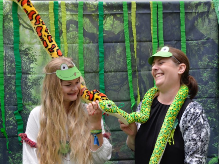 Zwei lachende junge Frauen. Beide tragen eine Papp-Frosch Maske auf der Stirn und Plüschschlangen auf den Schultern.