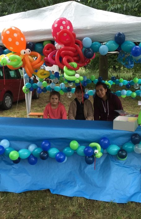 Junge Frau und zwei Kinder hinter einem Stand für Ballontiere. Der Stand ist mit einer roten und einer orangenen Ballonkrake, sowie blauen und grünen Ballons geschmückt.